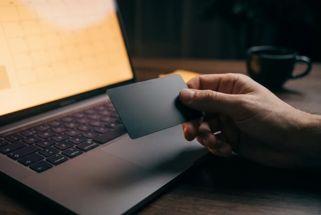 A close-up photograph captures a person's hand holding a sleek, blank credit card, with their index finger pausing in mid-air just above a laptop touchpad. The scene maintains the moody, low-key lighting established in the previous images, with deep shadows enveloping the background. A soft, warm amber glow from the slightly blurred laptop screen illuminates the hand, the card, and the touchpad, perfectly conveying the tense moment of hesitation before making a purchase decision. No text, numbers, or logos are visible on the credit card.
